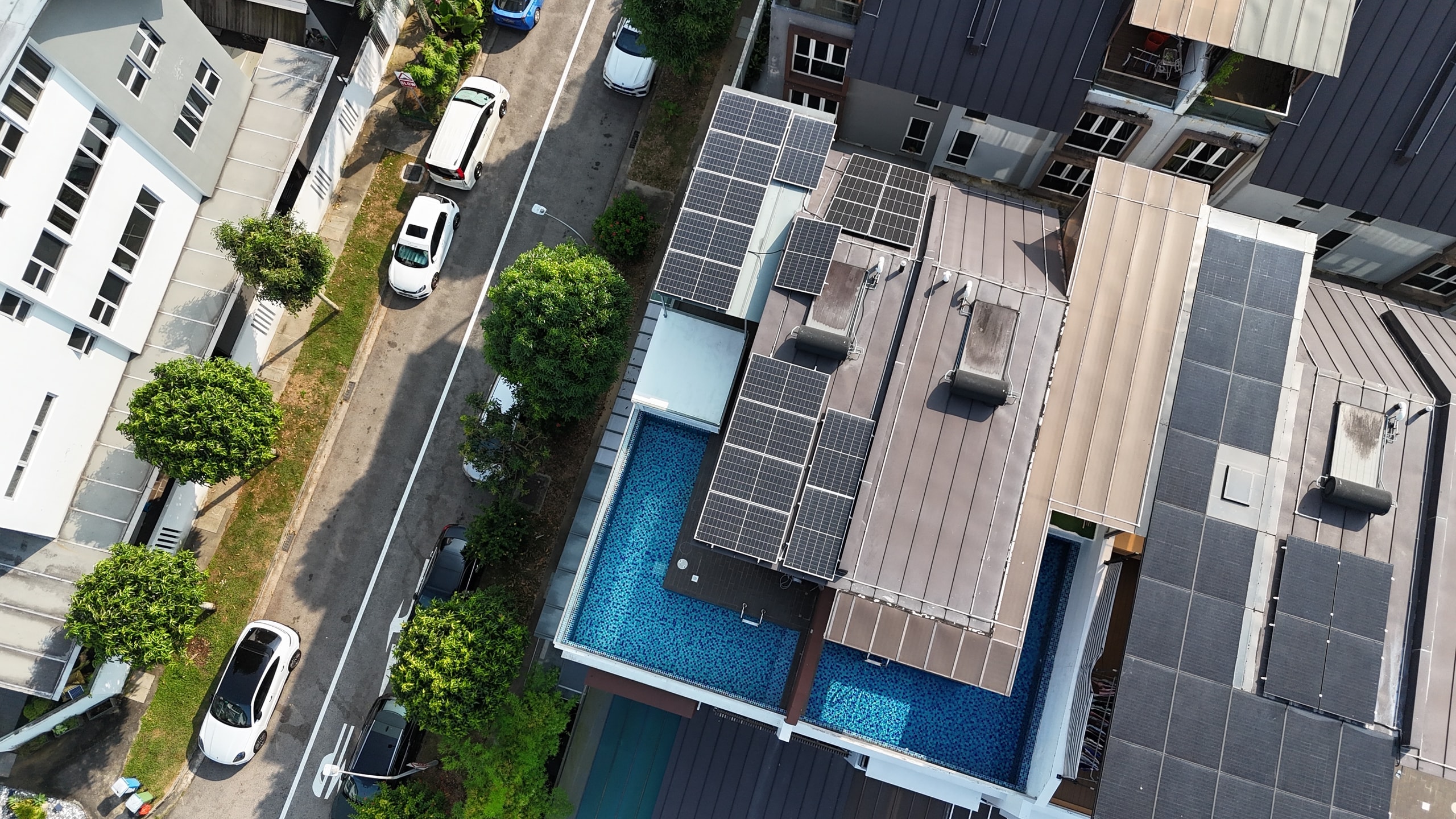 Drone view over a row of landed houses in Sembawang with pools, solar panels on the grey roofs, street and parked cars alongside
