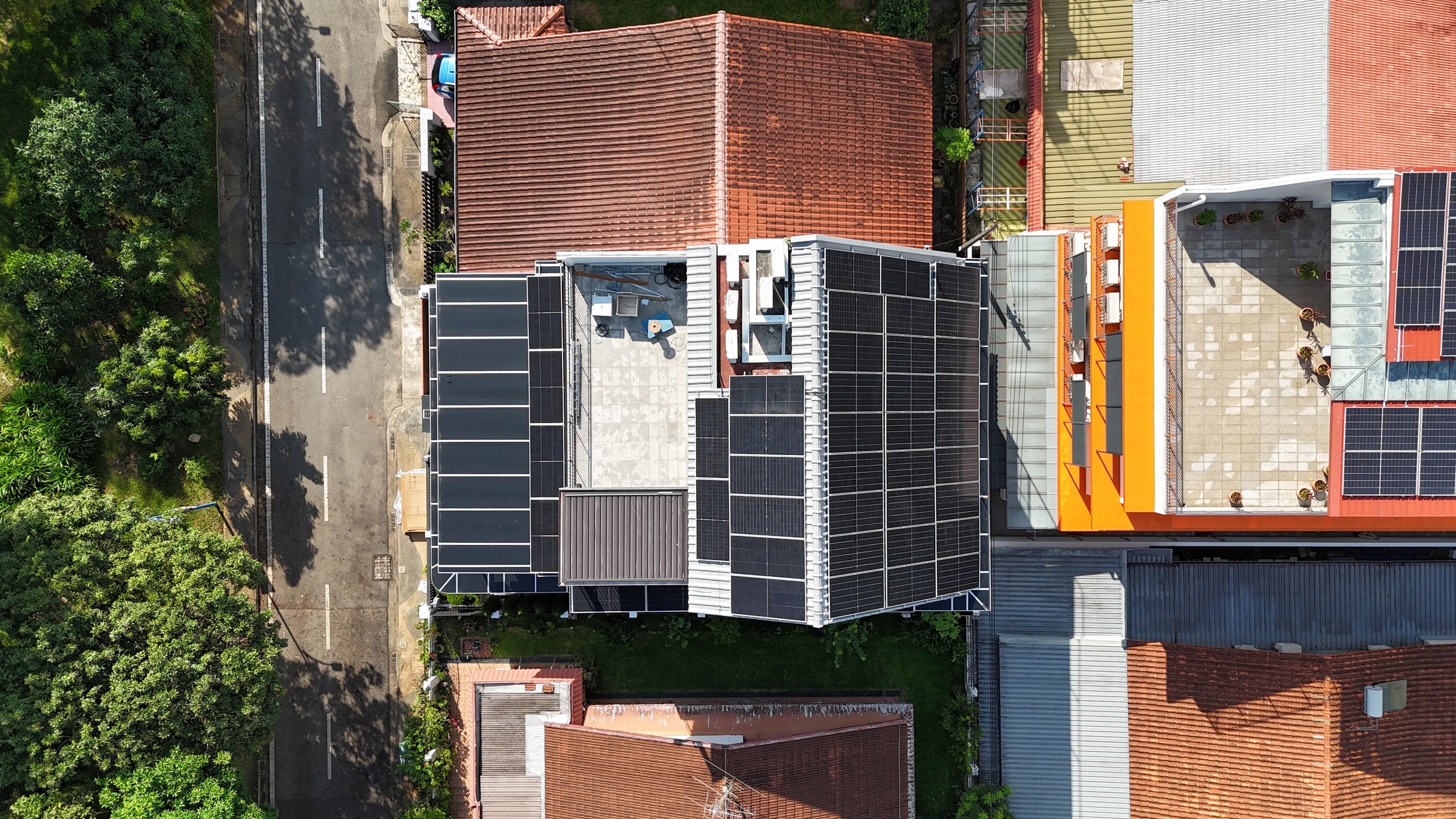 Drone view straight down onto a landed house in Kranji, panel rows across two grey roof sections beside a tree-lined road